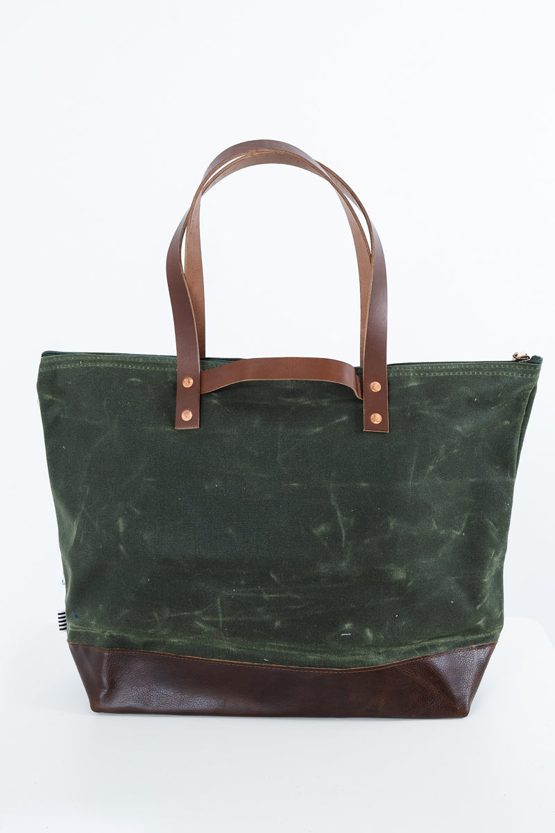 A green waxed canvas tote bag with dark brown leather handles and a matching dark brown leather base, shown against a plain white background. Handmade by Meanwhile Back on the Farm in Lynchburg,VA