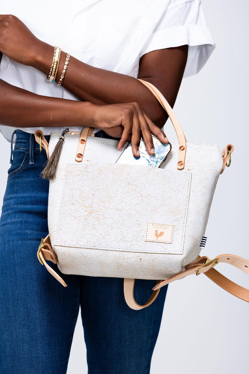 Picture of model placing her cell phone in the front pocket of a birch white leather purse with cream colored handles and back pack straps.
