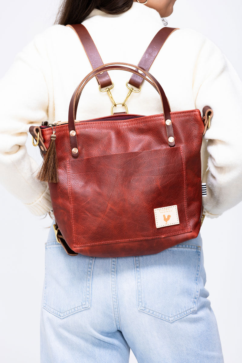 Model placing a ruby leather wallet into the front pocket of a medium sized ruby leather hand bag that she is holding by it's handles