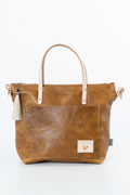 A brown textured leather tote bag with light tan handles, a front pocket, a white tassel charm, and a small brand patch on the bottom right, photographed on a white background. Handmade by Meanwhile Back on the Farm in Lynchburg,VA