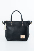 Black leather tote bag with dual top handles, a front slip pocket, a small cream leather patch with a rooster logo, and a white tassel charm on the left side, shown on a white background. Handmade by Meanwhile Back on the Farm in Lynchburg,VA