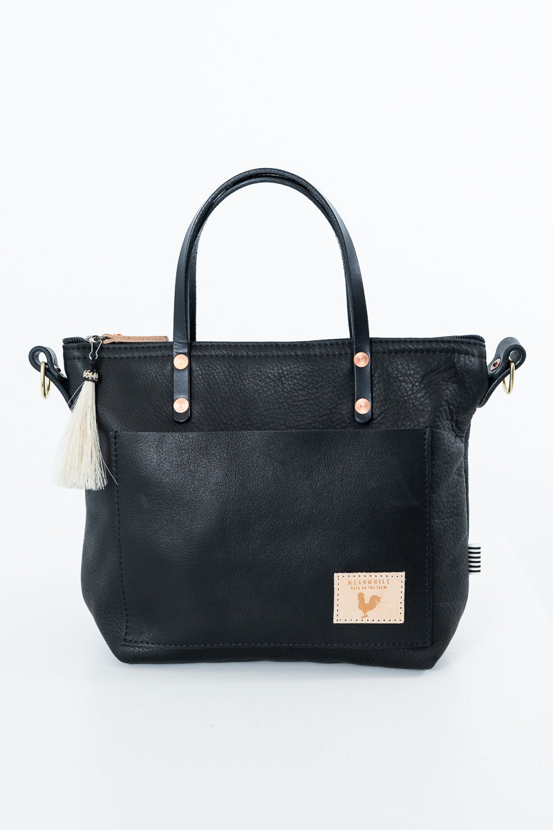 Black leather tote bag with dual top handles, a front slip pocket, a small cream leather patch with a rooster logo, and a white tassel charm on the left side, shown on a white background. Handmade by Meanwhile Back on the Farm in Lynchburg,VA