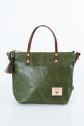 An olive green leather tote bag with brown leather handles, a front pocket, and a small rectangular brand patch on the lower right front, photographed on a clean white background. Handmade by Meanwhile Back on the Farm in Lynchburg,VA