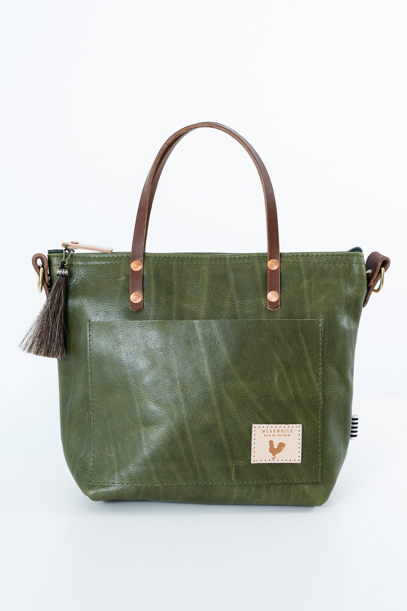 An olive green leather tote bag with brown leather handles, a front pocket, and a small rectangular brand patch on the lower right front, photographed on a clean white background. Handmade by Meanwhile Back on the Farm in Lynchburg,VA