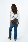 A woman viewed from behind wearing a white textured blouse, blue jeans, and brown shoes, carrying a dark brown leather handbag with gold-tone hardware over her shoulder against a plain white studio background. Handmade by Meanwhile Back on the Farm in Lynchburg,VA