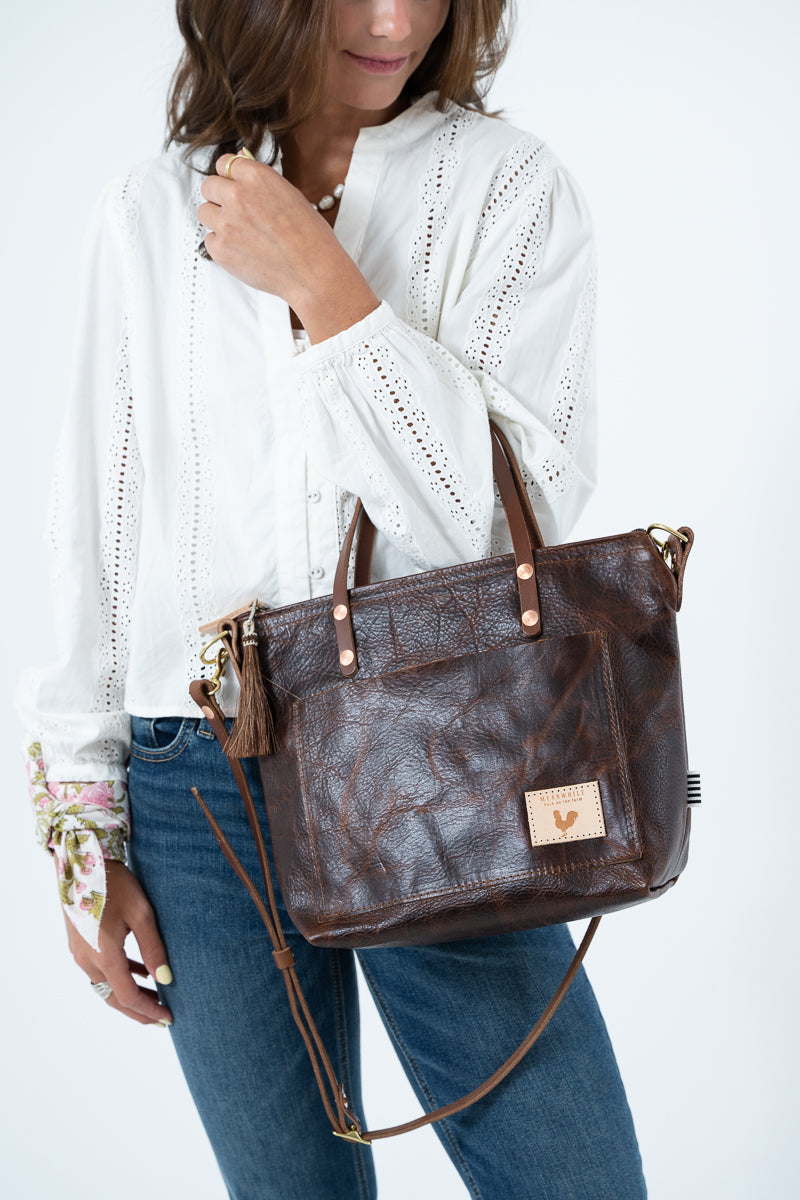A woman wearing a white blouse and blue jeans holds a dark brown leather tote bag with a floral fabric accent and a small gold-tone logo patch on the front, set against a plain white studio background. Handmade by Meanwhile Back on the Farm in Lynchburg,VA