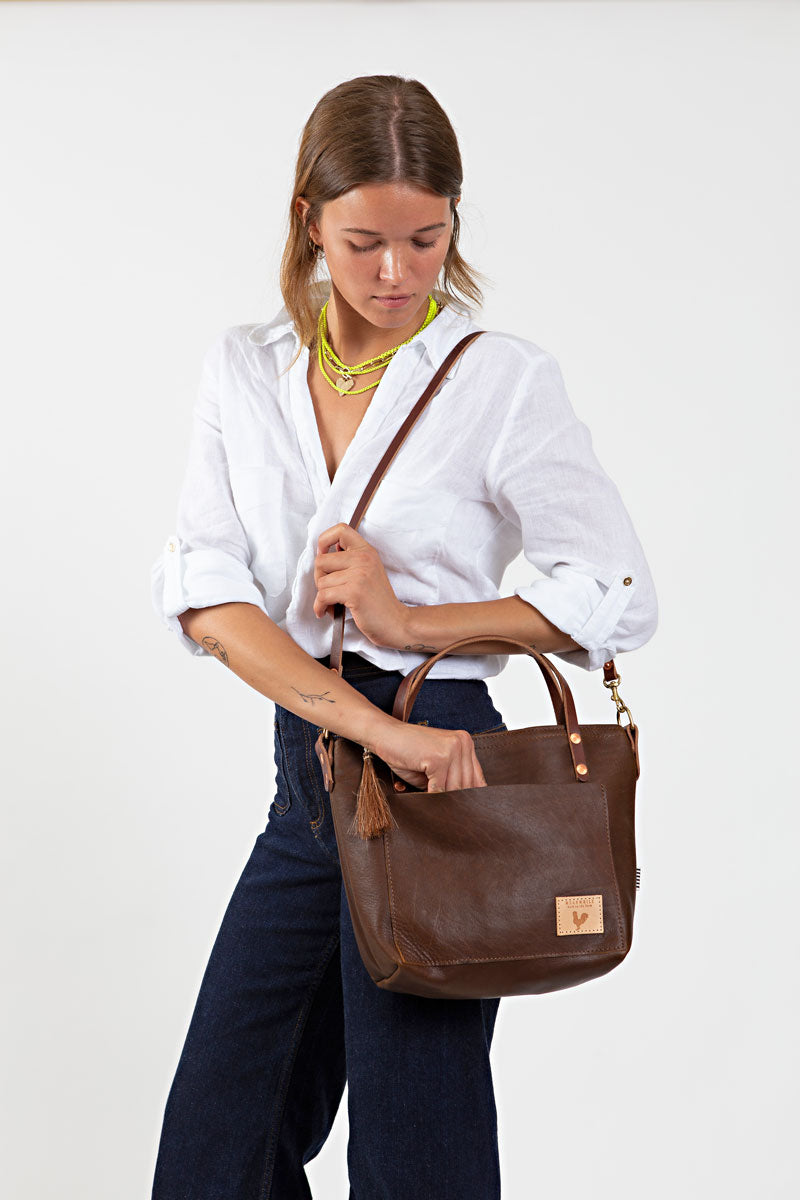 A woman holding a mocha brown short strapped bag with a brown tassel and meanwhile logo.