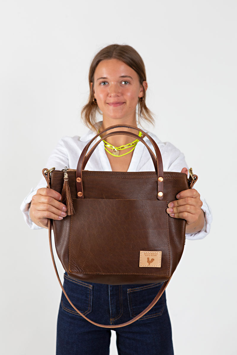 A woman holding the mocha brown bag with shoulder and cross body strap with meanwhile logo.