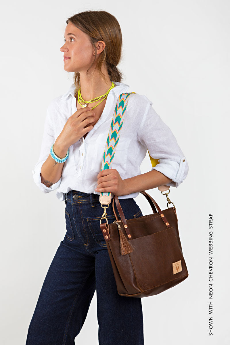 A woman holding a mocha brown bag with a green and orange design shoulder strap, a brown tassel, and meanwhile logo.