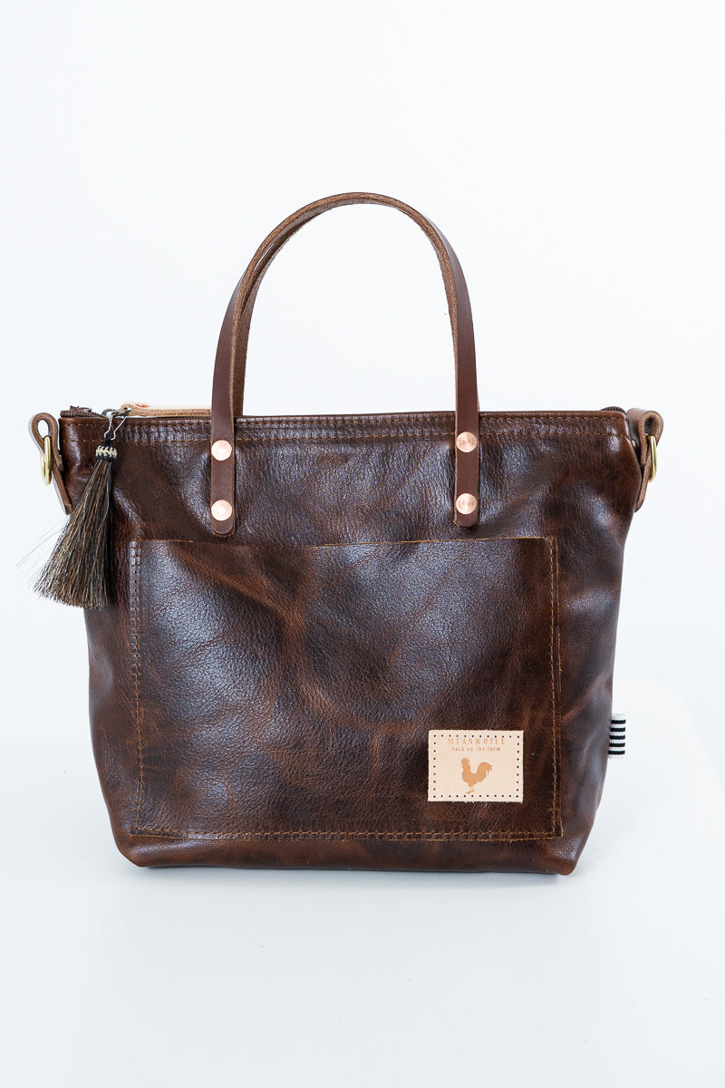 A woman holding a mocha brown short strapped bag with a brown tassel and meanwhile logo.