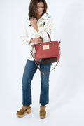 A woman modeling a dusty red and dark brown leather handbag with a gold-tone logo plaque, styled with a white embroidered shirt, blue jeans, and brown platform mule shoes against a white studio background. Handmade by Meanwhile Back on the Farm in Lynchburg,VA