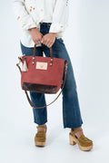 A person wearing a white embroidered blouse, blue jeans, and brown platform sandals holds a terracotta red waxed canvas tote bag with dark brown leather trim and handles. The bag features a small gold-tone logo patch on the front and a tassel detail on the side, set against a plain white studio background. Handmade by Meanwhile Back on the Farm in Lynchburg,VA