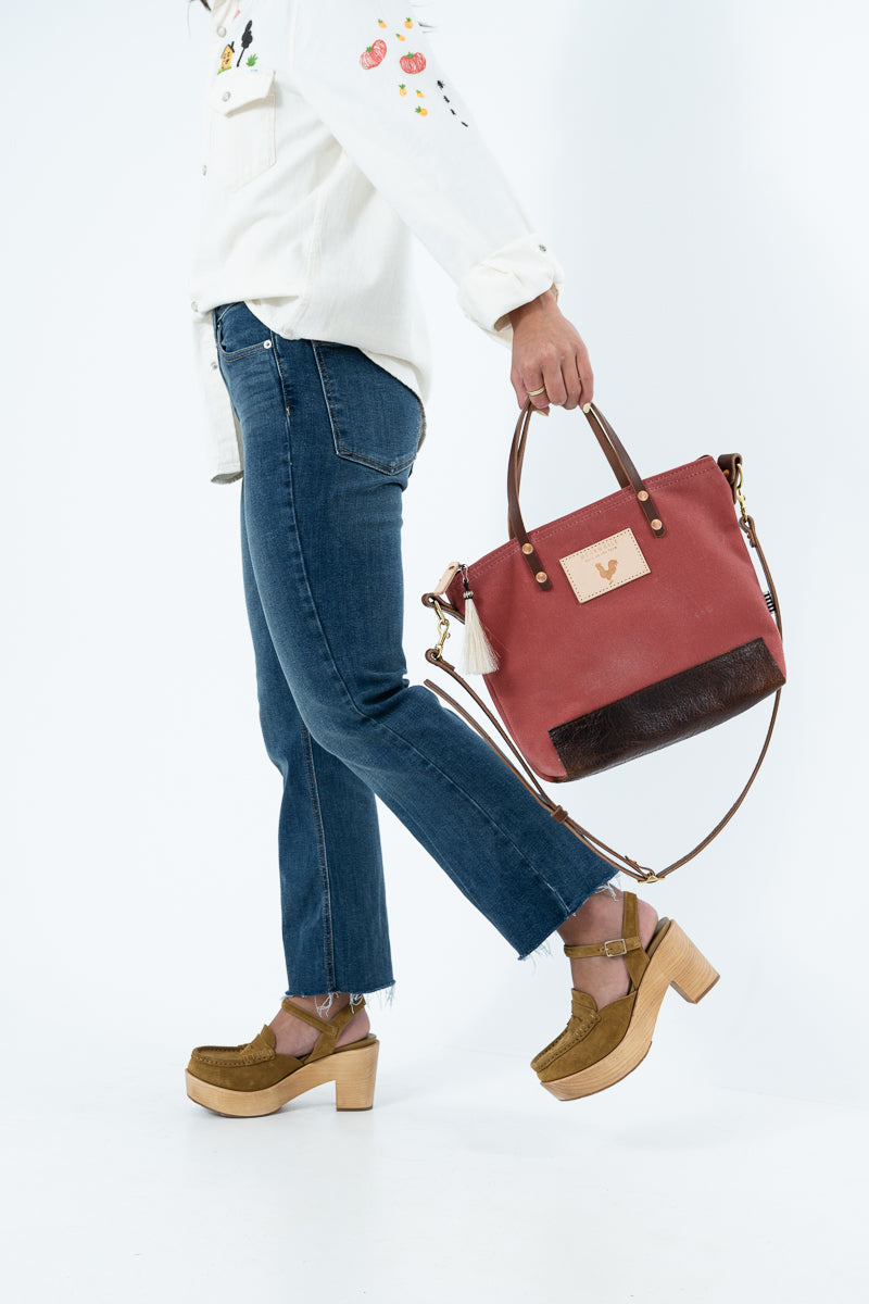A person wearing blue jeans and brown platform sandals carries a nantucket red waxed canvas tote bag with dark brown trim and a gold-tone logo plate on a white background.
