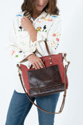 A woman wearing a white embroidered shirt and blue jeans carries a large reddish-brown waxed canvas tote bag with a dark brown front panel and gold-tone hardware against a plain white background. Handmade by Meanwhile Back on the Farm in Lynchburg,VA