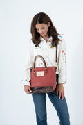 A woman wearing a white embroidered shirt and blue jeans holds a terracotta red waxed canvas tote bag with dark brown leather trim and handles against a plain white background. Handmade by Meanwhile Back on the Farm in Lynchburg,VA