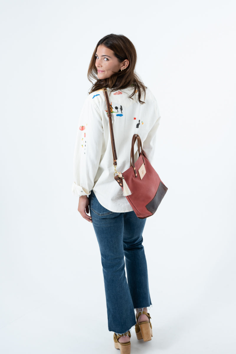 A woman wearing a white embroidered long-sleeve shirt, blue jeans, and tan wedge sandals, carrying a red waxed canvas shoulder bag with a brown leather bottom against a plain white studio background. Handmade by Meanwhile Back on the Farm in Lynchburg,VA
