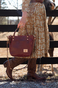 A person outdoors near a black wooden fence holds a burgundy leather handbag with a small tan logo patch. The person is wearing a beige and brown patterned midi dress and brown cowboy boots, with a horse partially visible behind the fence. Handcrafted by Meanwhile Back on the Farm in Lynchburg, Va.