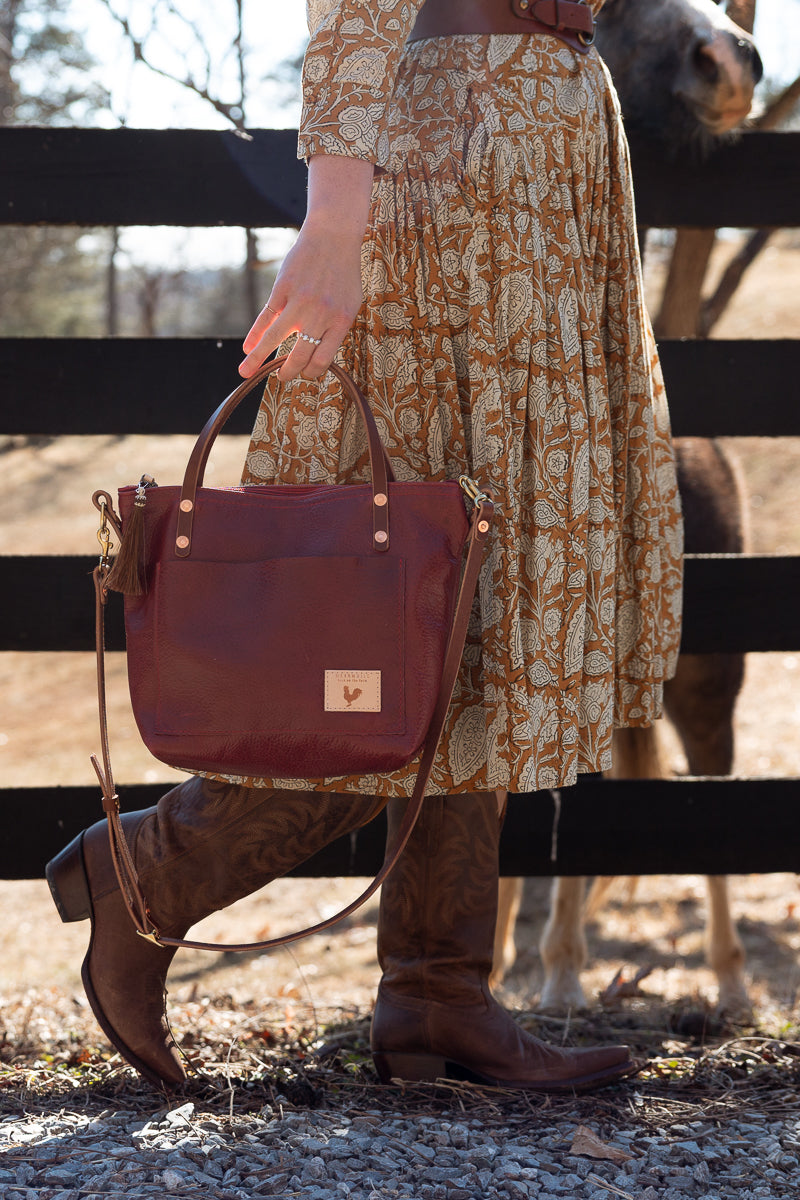 A person outdoors near a black wooden fence holds a burgundy leather handbag with a small tan logo patch. The person is wearing a beige and brown patterned midi dress and brown cowboy boots, with a horse partially visible behind the fence. Handcrafted by Meanwhile Back on the Farm in Lynchburg, Va.