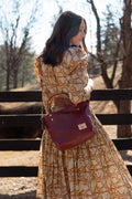 A woman wearing a long-sleeve beige and orange botanical print dress is shown from behind, carrying a dark reddish-brown leather shoulder bag with a gold logo plate. She is standing outdoors in front of a wooden fence with trees in the background. Handcrafted by Meanwhile Back on the Farm in Lynchburg, Va.