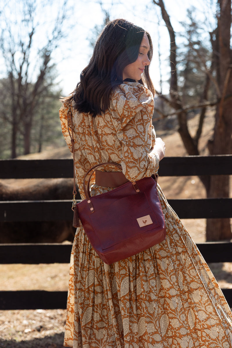 A woman wearing a long-sleeve beige and orange botanical print dress is shown from behind, carrying a dark reddish-brown leather shoulder bag with a gold logo plate. She is standing outdoors in front of a wooden fence with trees in the background. Handcrafted by Meanwhile Back on the Farm in Lynchburg, Va.