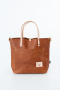 A brown leather tote bag with light tan handles and a front pocket, shown against a plain white background. Handmade by Meanwhile Back on the Farm in Lynchburg,VA