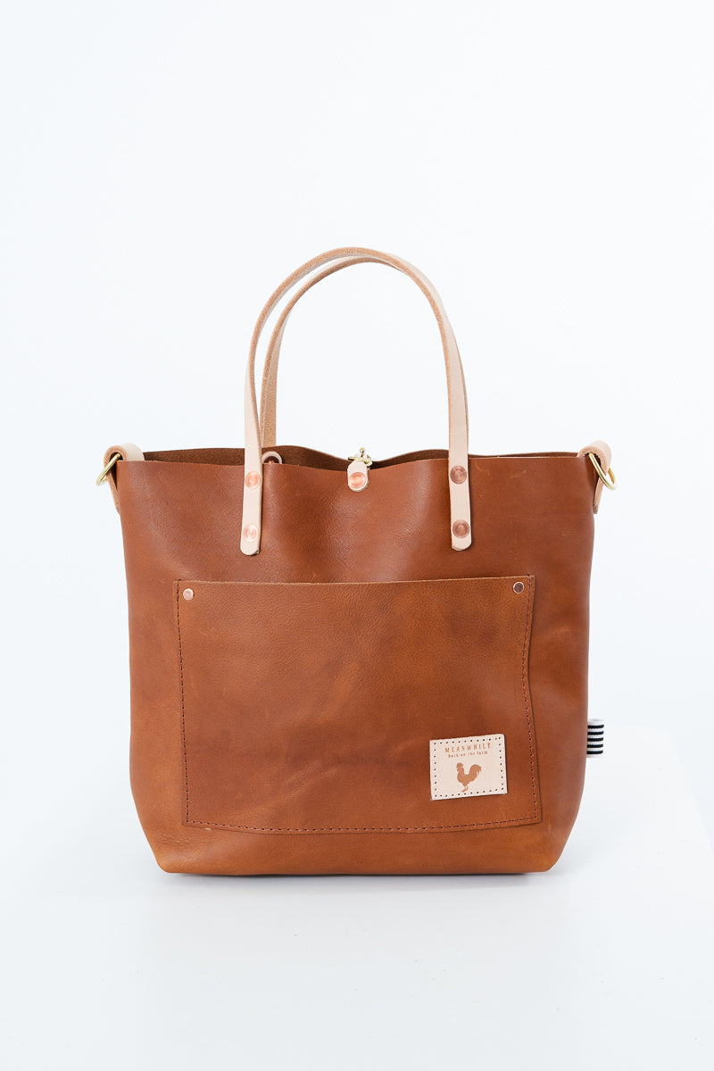 A brown leather tote bag with light tan handles and a front pocket, shown against a plain white background. Handmade by Meanwhile Back on the Farm in Lynchburg,VA