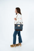 A woman in a white embroidered shirt, blue jeans, and brown platform shoes is shown from the side, carrying a navy blue tote bag with brown leather trim and a small gold logo patch on a white studio background. Handmade by Meanwhile Back on the Farm in Lynchburg,VA