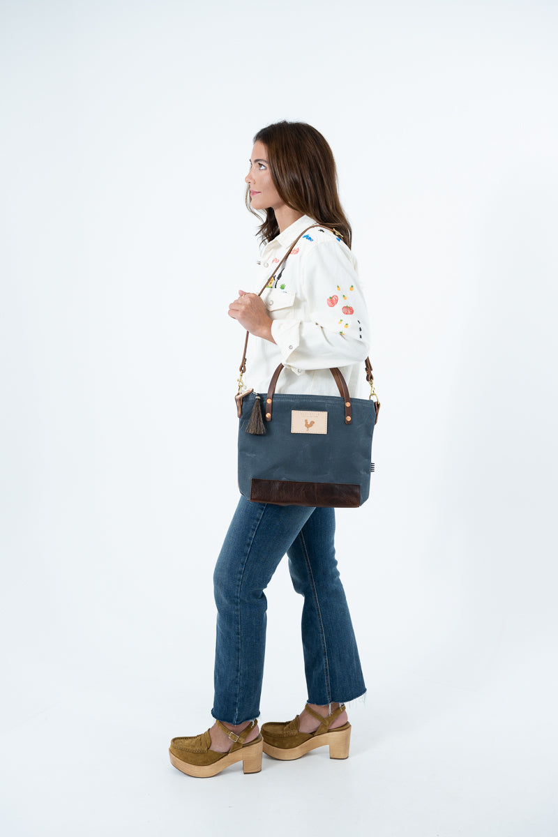 A woman in a white embroidered shirt, blue jeans, and brown platform shoes is shown from the side, carrying a navy blue tote bag with brown leather trim and a small gold logo patch on a white studio background. Handmade by Meanwhile Back on the Farm in Lynchburg,VA