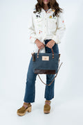 A model wearing a white embroidered shirt, blue jeans, and brown platform shoes, holding a slate blue tote bag with dark brown leather trim and handles. Handmade by Meanwhile Back on the Farm in Lynchburg,VA