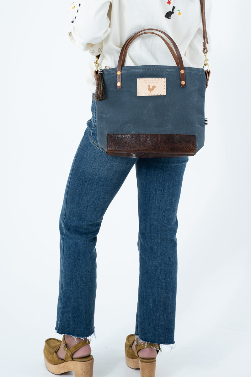 A woman wearing a white embroidered button-up shirt and blue jeans carries a navy blue waxed canvas tote bag with brown leather handles and a brown leather bottom panel. The bag features a tan patch with a rooster logo and a tassel on the side, set against a plain white background. Handmade by Meanwhile Back on the Farm in Lynchburg,VA