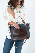 A woman wearing a white embroidered button-up shirt and blue jeans carries a large leather shoulder bag with a dark blue body and a brown front pocket and straps, photographed against a plain white background. Handmade by Meanwhile Back on the Farm in Lynchburg,VA