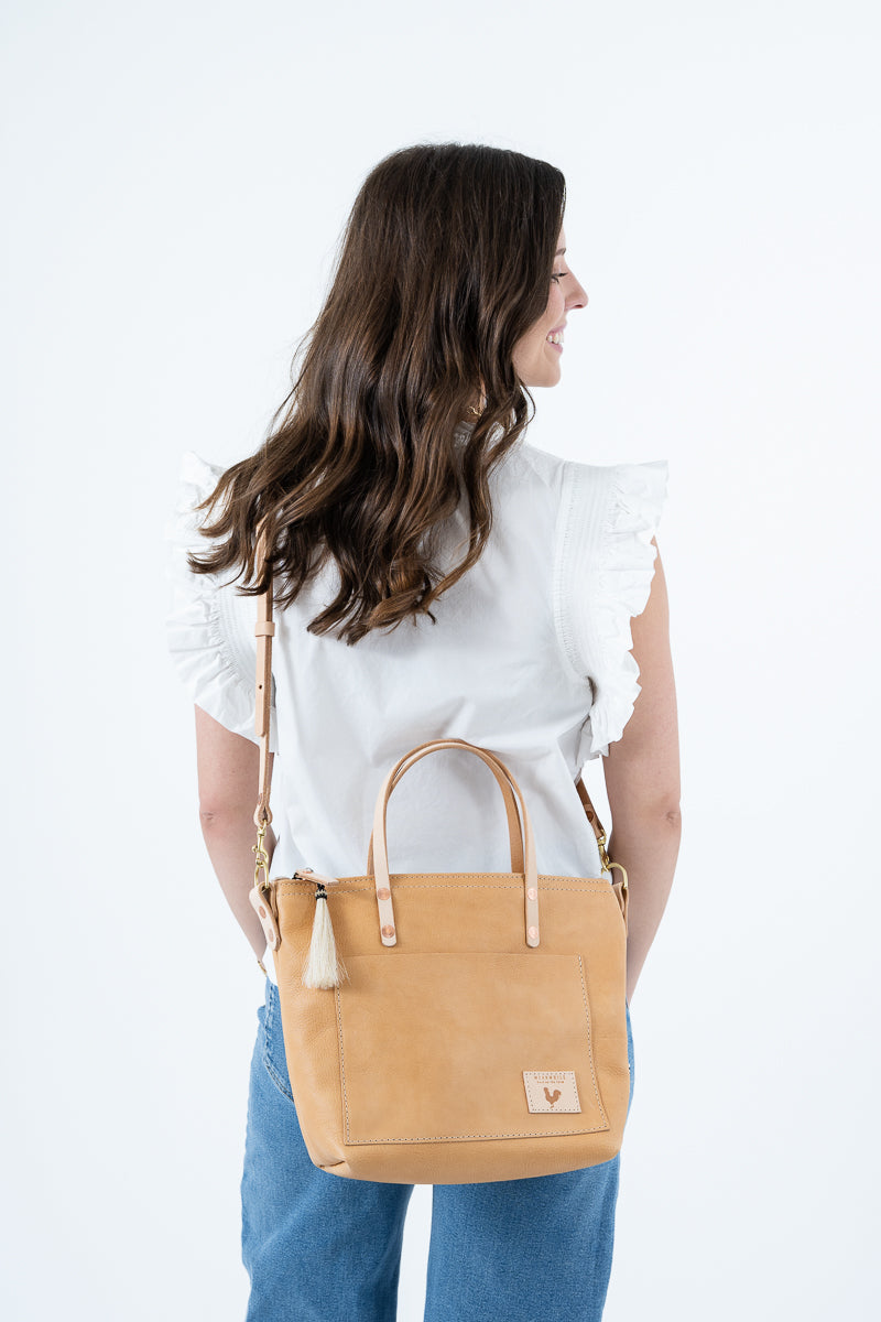 A woman with long brown hair is shown from behind wearing a white ruffled top and blue jeans, carrying a tan leather tote bag with a shoulder strap and tassel detail against a plain white background. Handmade by Meanwhile Back on the Farm in Lynchburg,VA