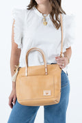 A woman wearing a white ruffled-sleeve blouse and blue jeans carries a tan leather tote bag with top handles and a shoulder strap against a plain white background. Handmade by Meanwhile Back on the Farm in Lynchburg,VA