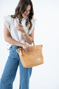 A woman wearing a white ruffled sleeveless top and blue jeans holds a tan leather tote bag with a small rooster shaped logo patch on the front pocket, set against a plain white background. Handmade by Meanwhile Back on the Farm in Lynchburg,VA