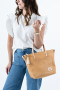 A woman wearing a white ruffled blouse and blue jeans carries a tan leather tote bag with cream tassel and trim against a white studio background. Handmade by Meanwhile Back on the Farm in Lynchburg,VA