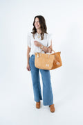 A woman stands against a white studio background wearing a white ruffled short-sleeve top, blue wide-leg jeans, and brown boots, while carrying two tan leather tote bags. One leather tote bag is new and the other has a beautifully worn patina. Handmade by Meanwhile Back on the Farm in Lynchburg,VA