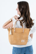 A woman in a white ruffled blouse and blue jeans holds a tan leather tote bag with light brown leather handles and a small leather brand patch on the front pocket, photographed against a clean white background. Handmade by Meanwhile Back on the Farm in Lynchburg,VA