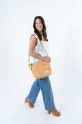 A woman wearing a white sleeveless top, blue jeans, and brown clogs carries a tan leather shoulder bag with a small square logo patch on a plain white studio background. Handmade by Meanwhile Back on the Farm in Lynchburg,VA