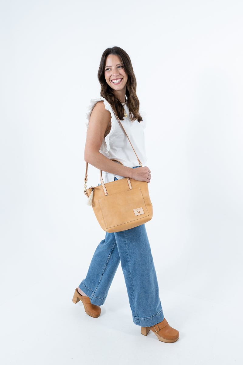 A woman wearing a white sleeveless top, blue jeans, and brown clogs carries a tan leather shoulder bag with a small square logo patch on a plain white studio background. Handmade by Meanwhile Back on the Farm in Lynchburg,VA
