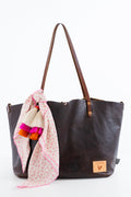 A dark brown leather tote bag with brown shoulder straps, featuring a light pink patterned scarf with colorful tassels draped over one side, set against a plain white background.