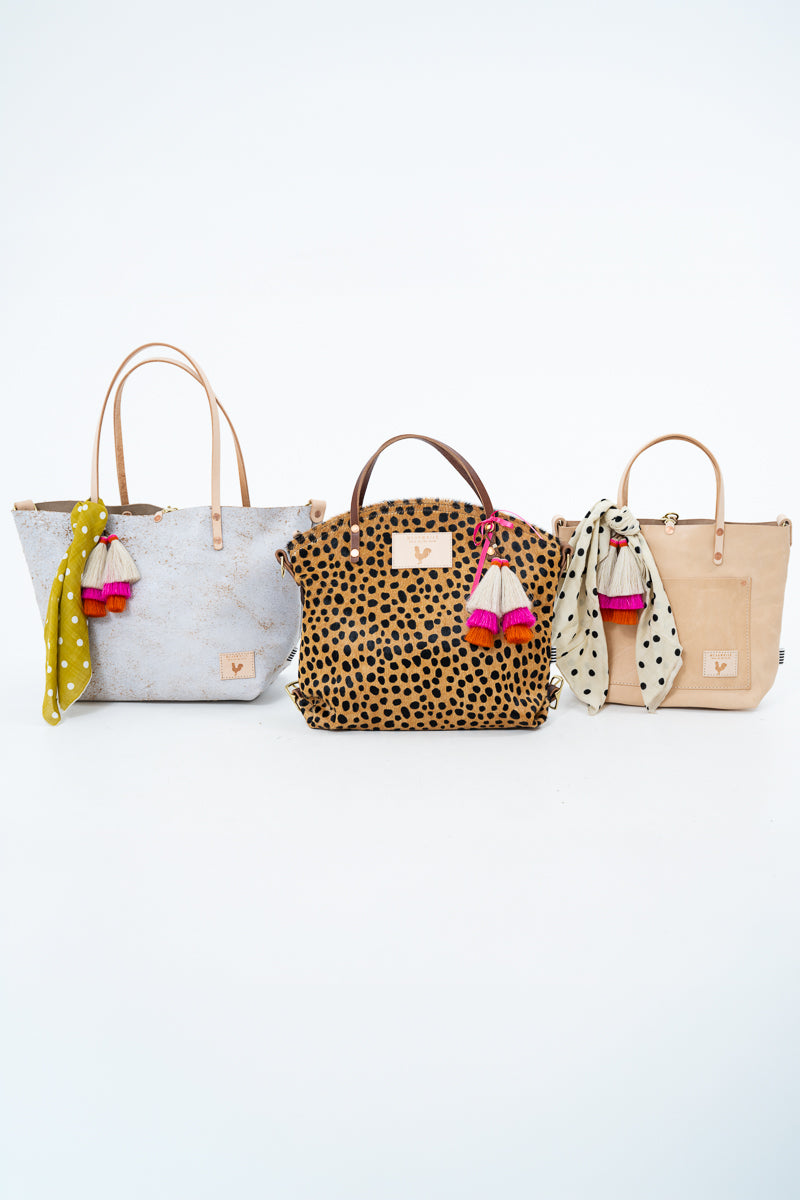 Three tote bags lined up against a white background: a distressed light gray bag with tan handles and a yellow polka-dot scarf charm, a leopard-print bag with brown handles and a pink-and-white scarf charm, and a beige bag with tan handles and a black-and-white polka-dot scarf charm. Handmade by Meanwhile Back on the Farm in Lynchburg,VA