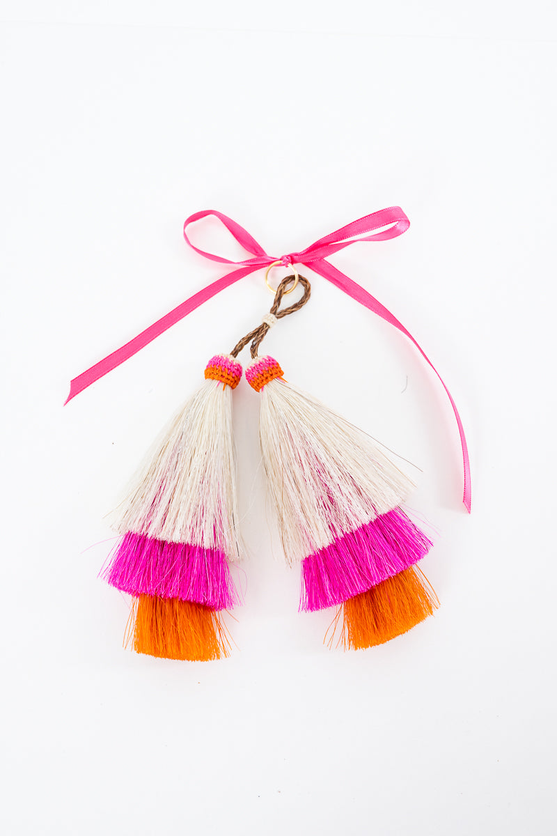 A close-up of a leopard-print handbag with brown leather trim and a hanging tassel charm. The charm has three tiers: cream, hot pink, and orange, tied to a brown strap with a pink ribbon.