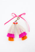 A pair of handmade tassel earrings with cream-colored raffia fibers, vibrant pink and orange bands, and a pink ribbon tied above them on a white background. Handmade by Meanwhile Back on the Farm in Lynchburg,VA