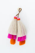 A pair of handmade tassels featuring cream-colored fibers, bright pink and orange fringe accents, and a brown cord with a gold ring, shown on a plain white background. Handmade by Meanwhile Back on the Farm in Lynchburg,VA