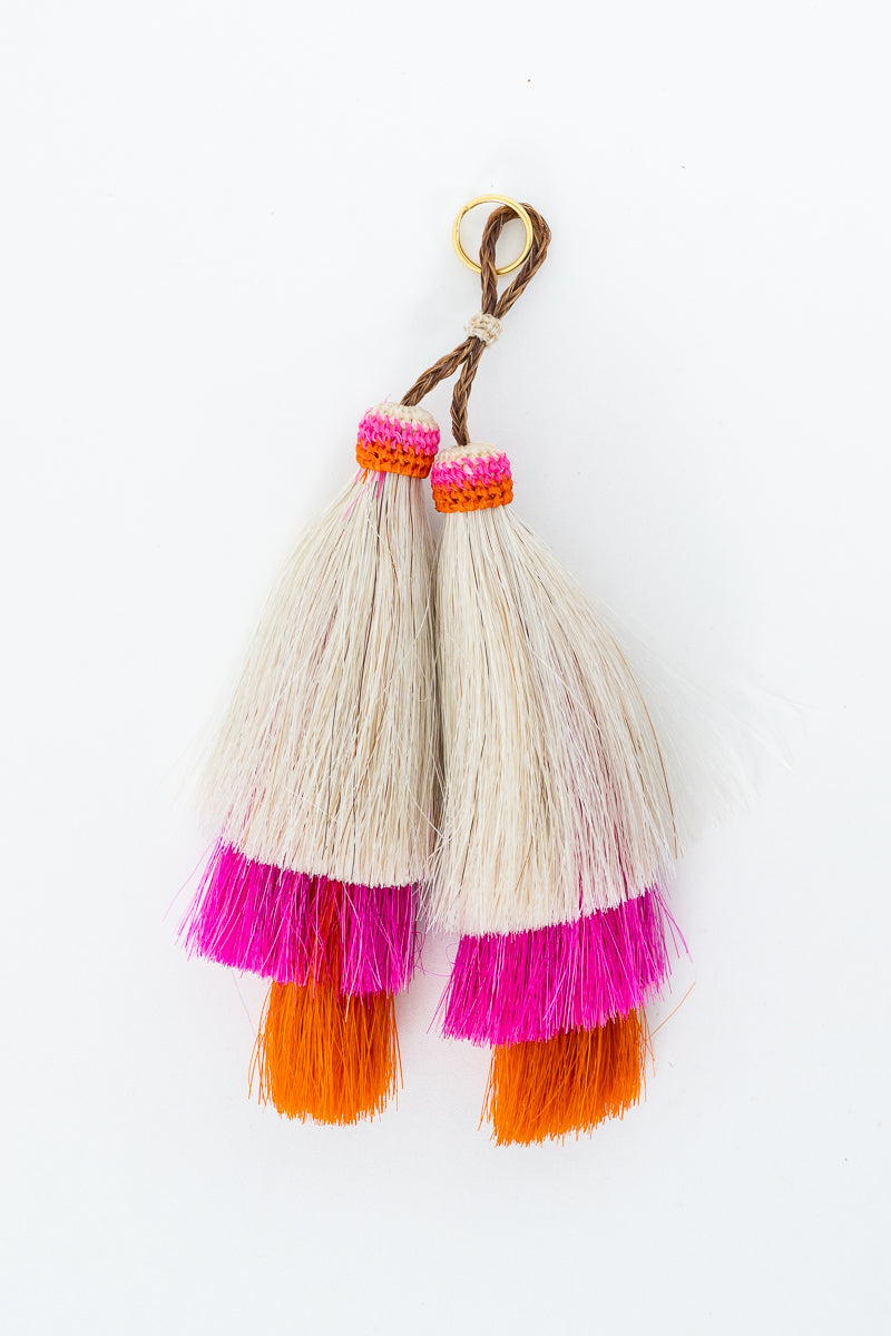 A pair of handmade tassels featuring cream-colored fibers, bright pink and orange fringe accents, and a brown cord with a gold ring, shown on a plain white background. Handmade by Meanwhile Back on the Farm in Lynchburg,VA