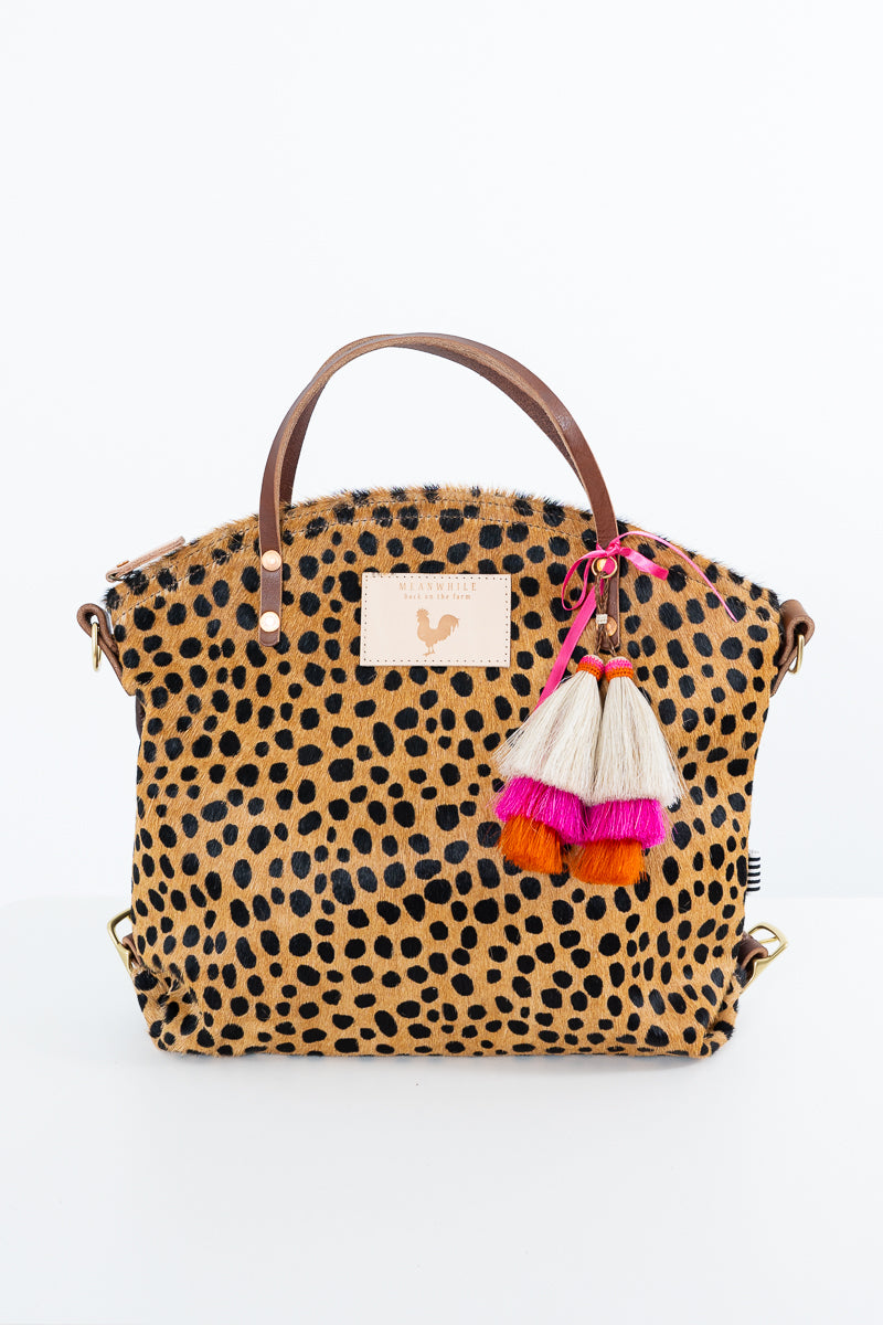 A leopard print handbag with brown leather handles, a cream brand patch, and a decorative tassel charm with pink, orange, and cream tassels, shown on a white background. Handmade by Meanwhile Back on the Farm in Lynchburg,VA