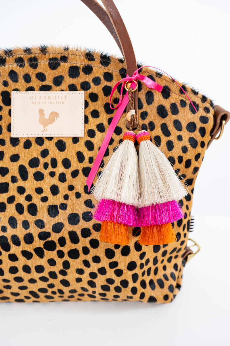 A close-up of a leopard-print handbag with brown leather trim and a hanging tassel charm. The charm has three tiers: cream, hot pink, and orange, tied to a brown strap with a pink ribbon.