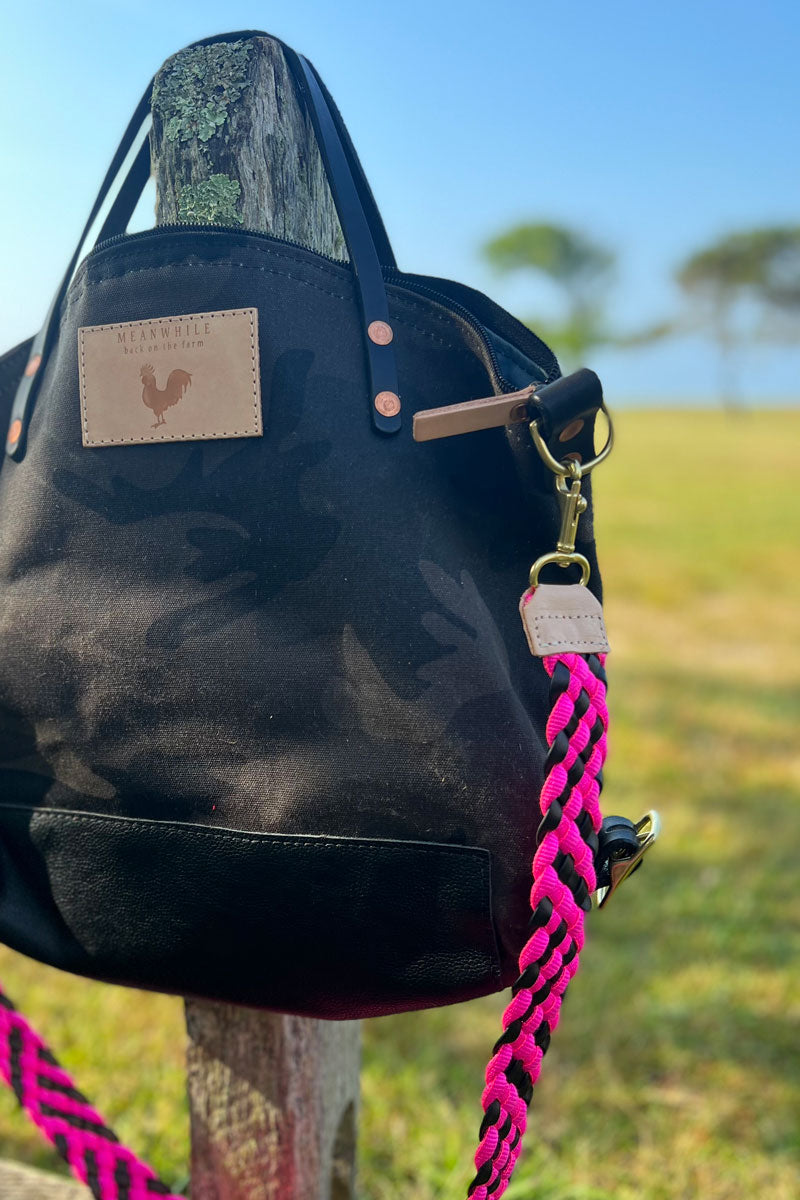 Black wax canvas camo handbag with a bright pink woven crossbody strap on fencepost