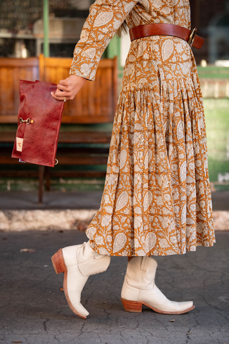 Woman in orange dress wearing white cowgirl boots holding a red/brown leather fold-over clutch and crossbody behind her