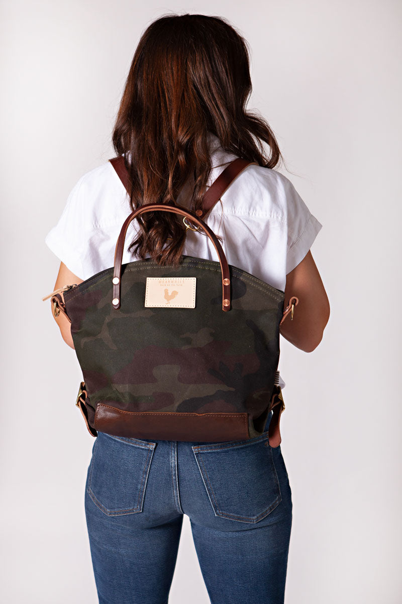 A woman wearing the camouflage backpack with dark brown straps and the meanwhile logo.